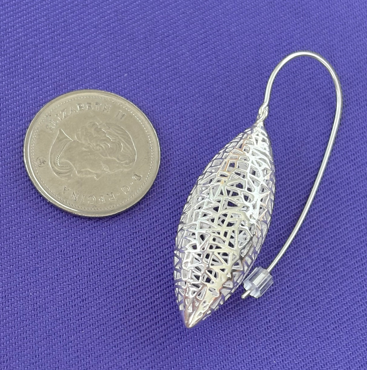 Silver pod shaped earring next to a coin on a purple background