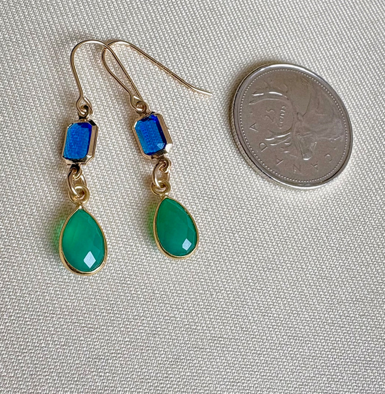 Gold earrings with blue and green teardrop stones next to a coin on a textured fabric background