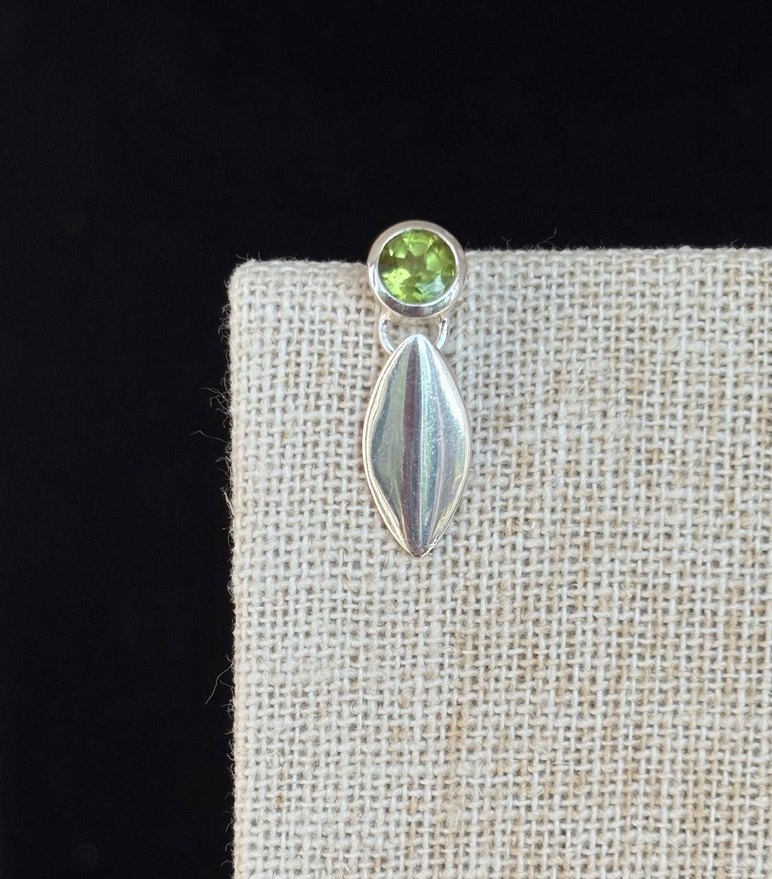 Peridot gemstone stud earring with a silver leaf drop on a textured beige surface