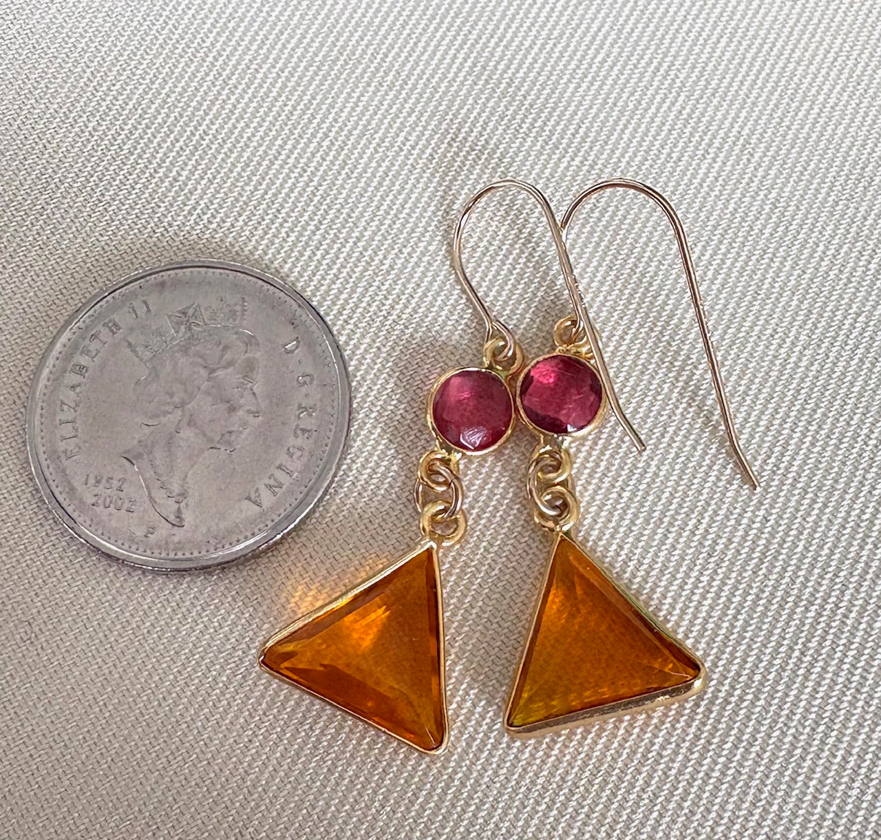 Pair of earrings with orange triangle drops and pink gemstones next to a quarter on a textured surface.