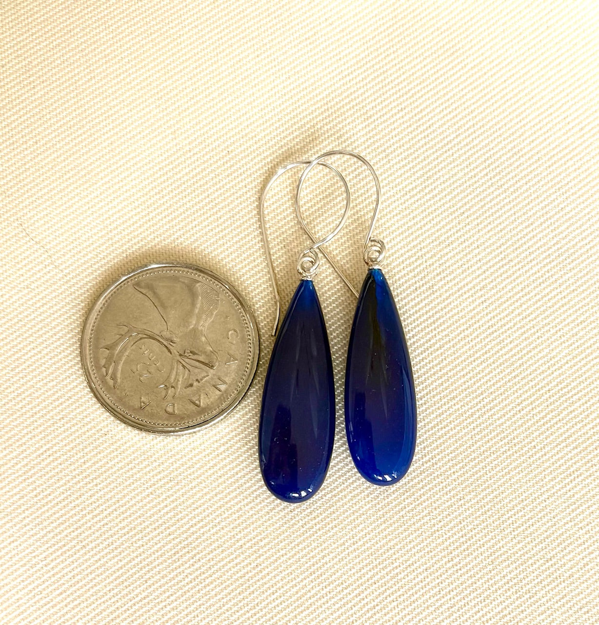 Blue teardrop earrings next to a quarter on a beige fabric background