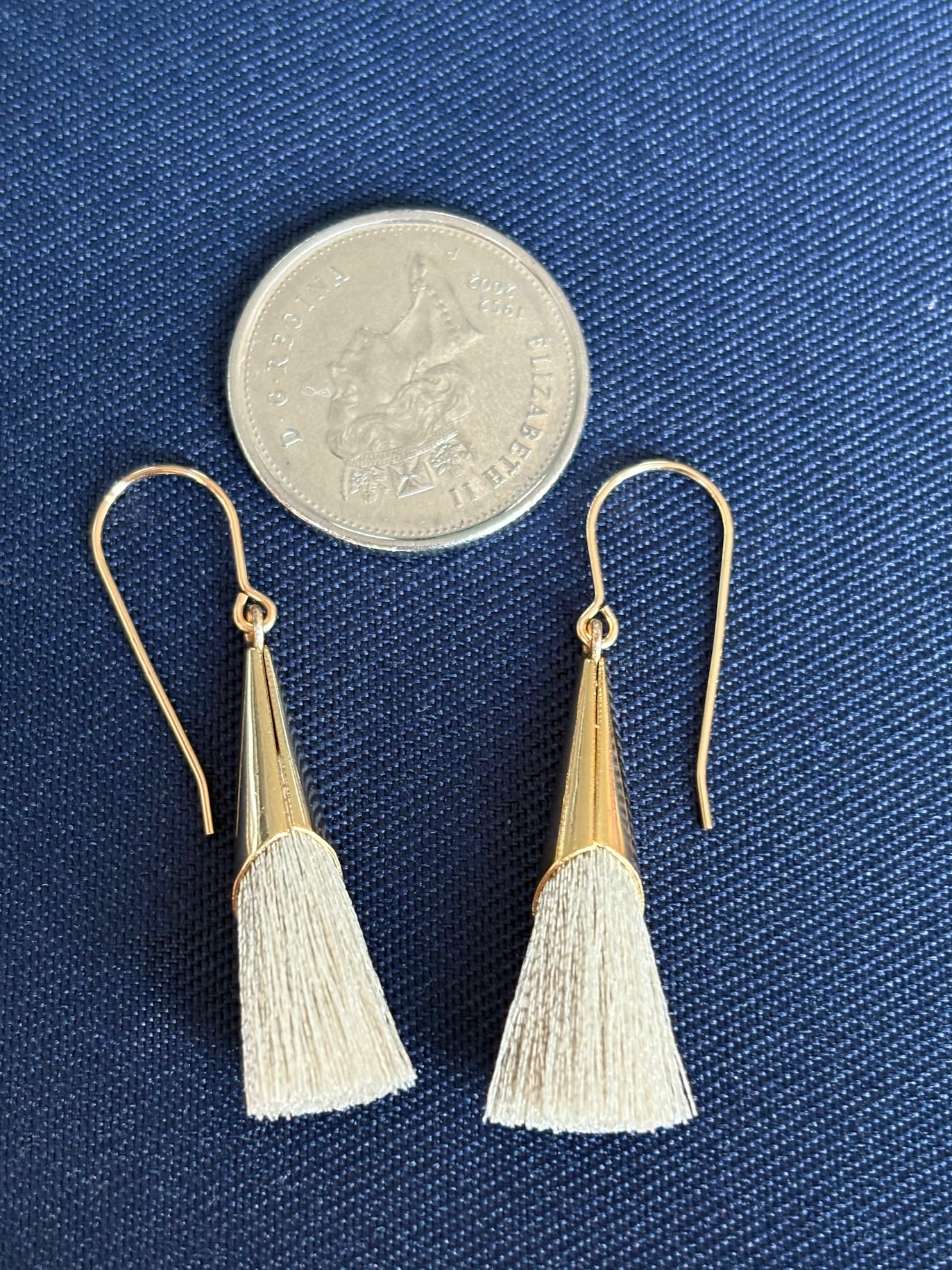 Gold and beige tassel earrings on a blue fabric background with a coin for scale.
