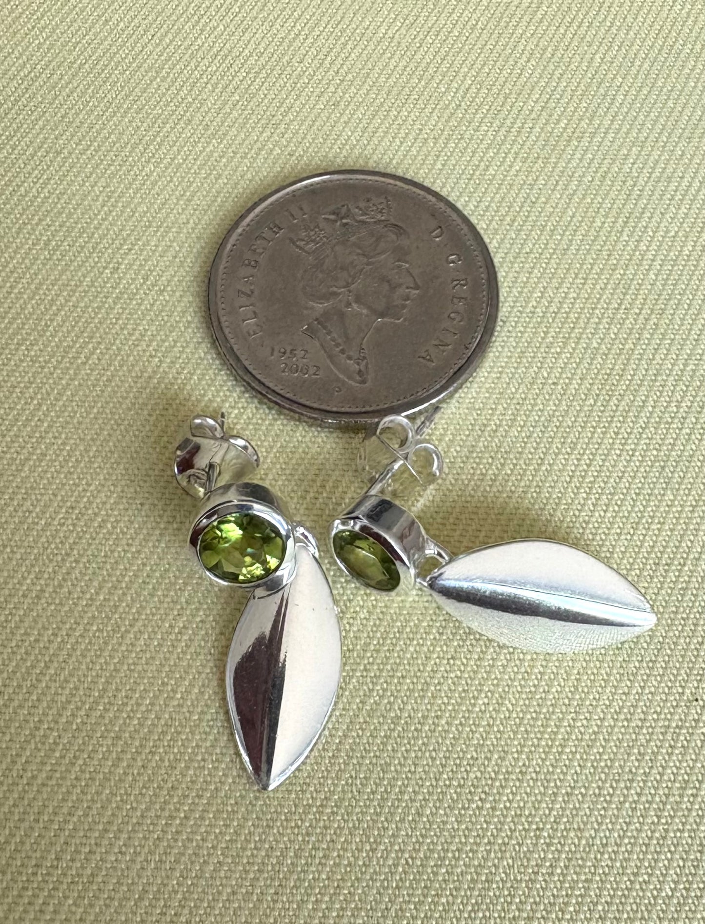 Silver leaf-shaped earrings with green gemstones next to a coin on a textured surface