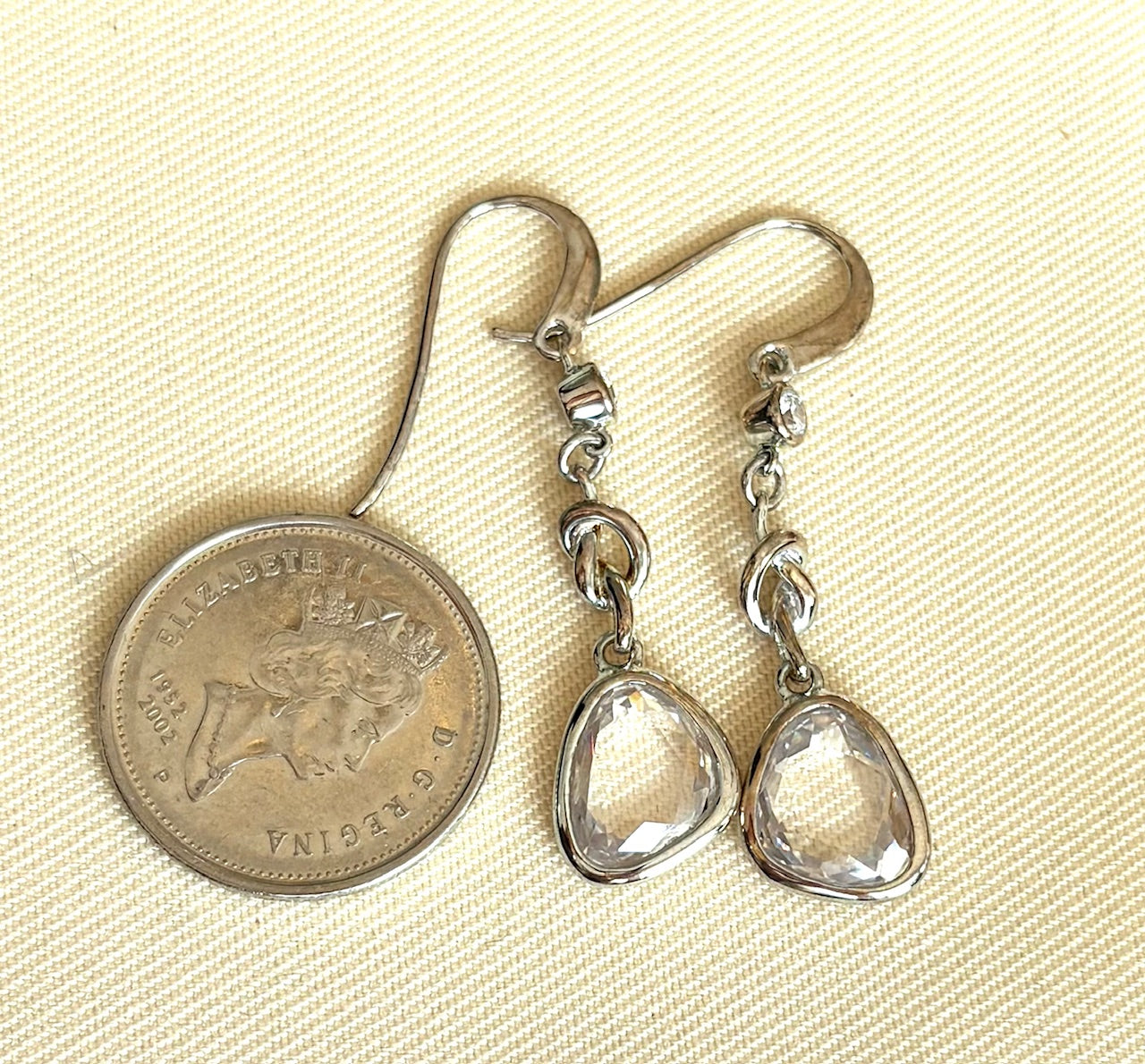 Teardrop-shaped silver earrings on a beige background with a coin for scale.