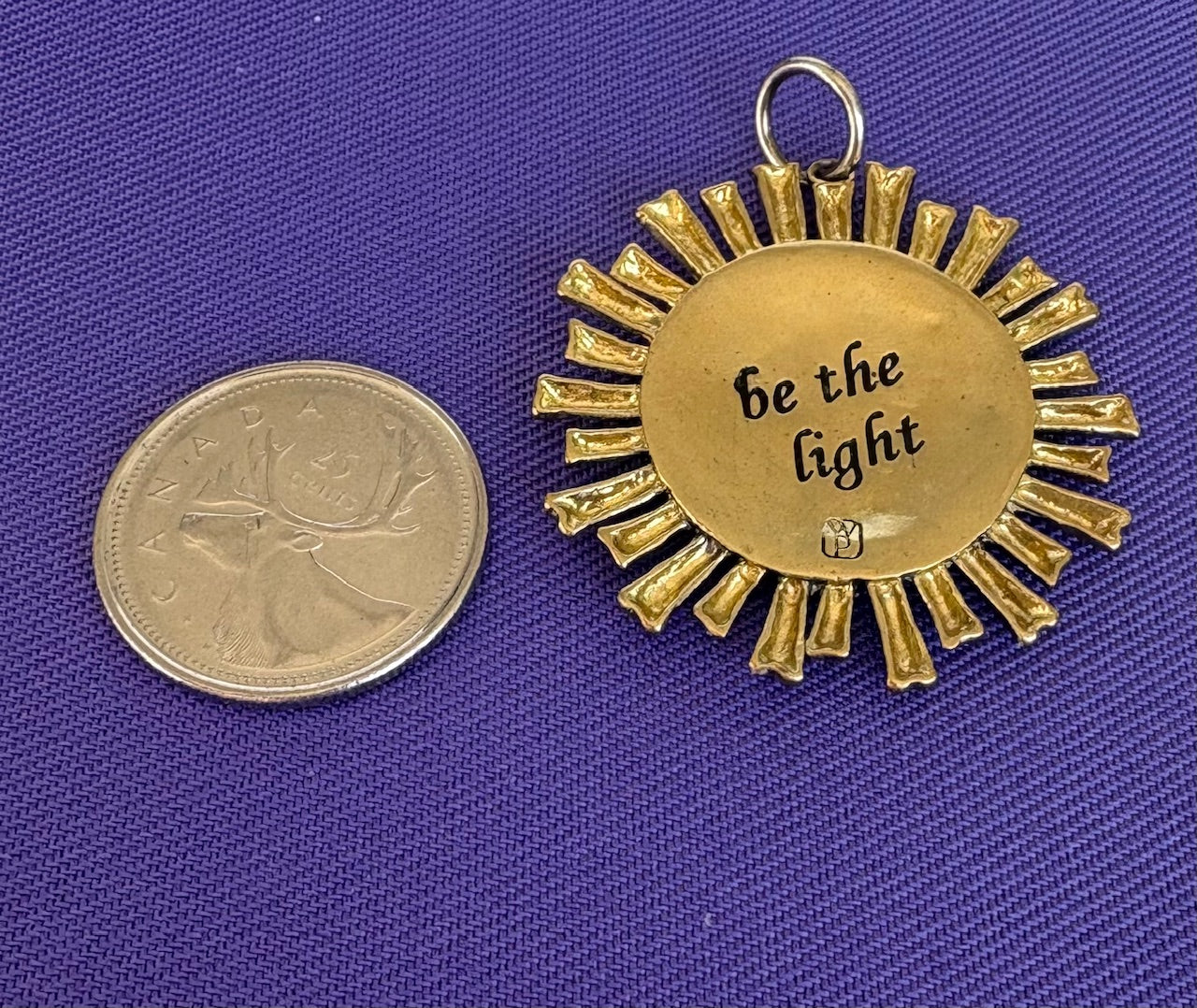 Gold sun-shaped pendant with 'be the light' text next to a coin on a blue background. Made by Waxing Poetic