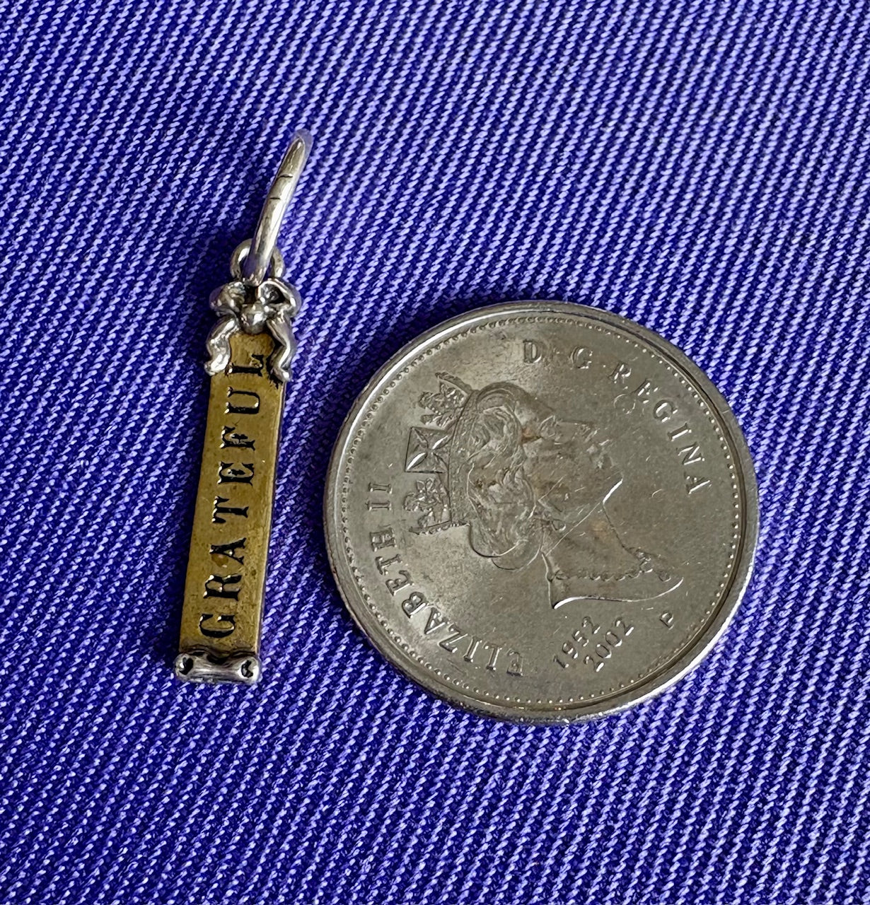 Small silver and gold 'GRATEFUL' pendant next to a coin on a blue textured background