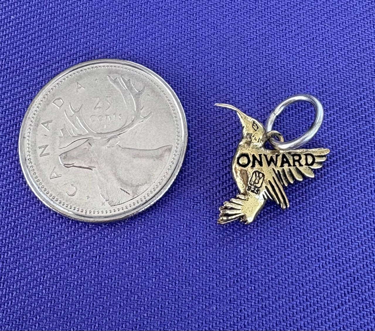 Small sterling silver hummingbird charm with 'ONWARD' text next to a Canadian quarter on a blue background