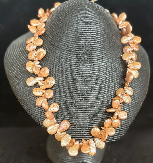 Necklace made of peach & bronze coloured keshi petal pearls on a gray stand against a dark background